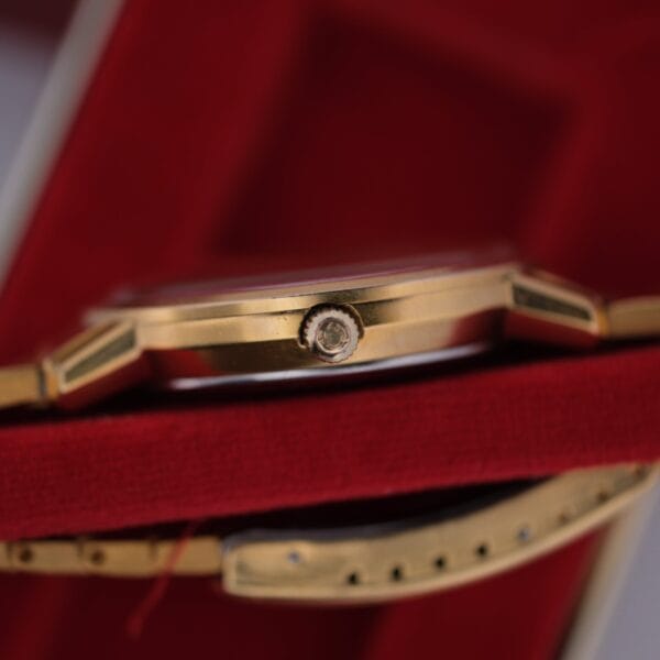 Close-up of an Omega Deville Automatic Gold Tone Date Full Set watch on red velvet, highlighting its side profile. The gold-tone case and detailed crown are prominent, with a glimpse of the gold wristband underneath.
