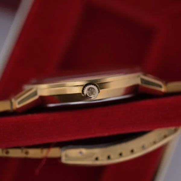 Close-up of an Omega Deville Automatic Gold Tone Date Full Set watch on red velvet, highlighting its side profile. The gold-tone case and detailed crown are prominent, with a glimpse of the gold wristband underneath.