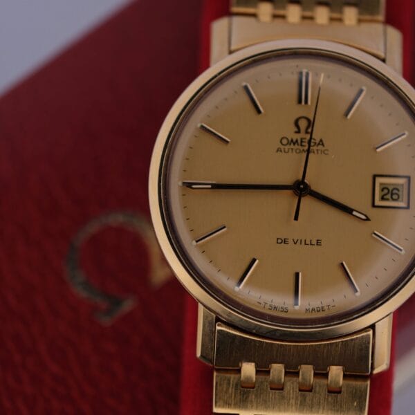 An Omega Deville Automatic Gold Tone wristwatch, featuring a date display on the right side, elegantly rests on its textured red case with a matching gold bracelet.