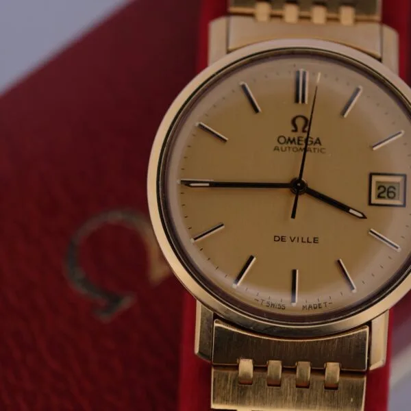 An Omega Deville Automatic Gold Tone wristwatch, featuring a date display on the right side, elegantly rests on its textured red case with a matching gold bracelet.