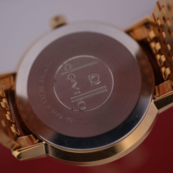The Omega Deville Automatic Gold Tone Date Full Set wristwatch features a metal bracelet and an engraved gold back with the brand logo and circular design, elegantly resting on a red and white surface.