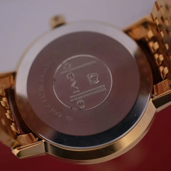 The Omega Deville Automatic Gold Tone Date Full Set wristwatch features a metal bracelet and an engraved gold back with the brand logo and circular design, elegantly resting on a red and white surface.