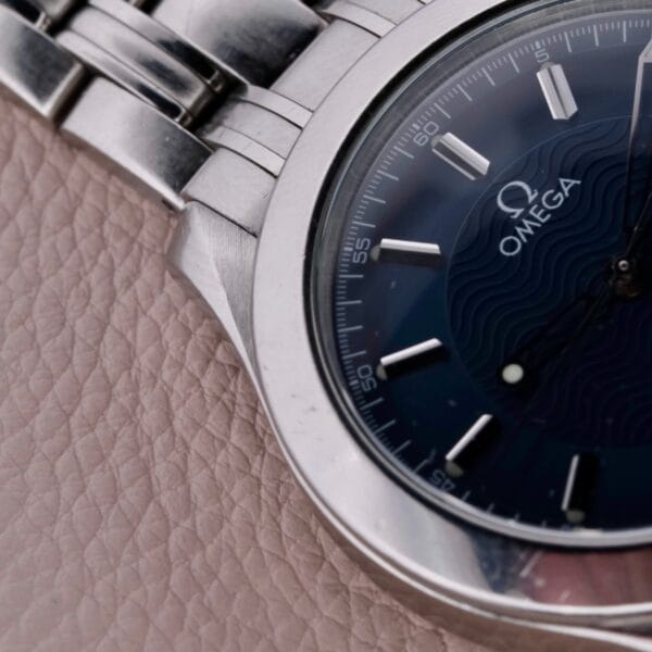 A close-up of the Omega Seamaster 120M Blue Wave shows its stainless steel band on a textured beige surface, featuring a dark blue face with white hour markers and Omega branding, all encased within a sleek, polished metal case.