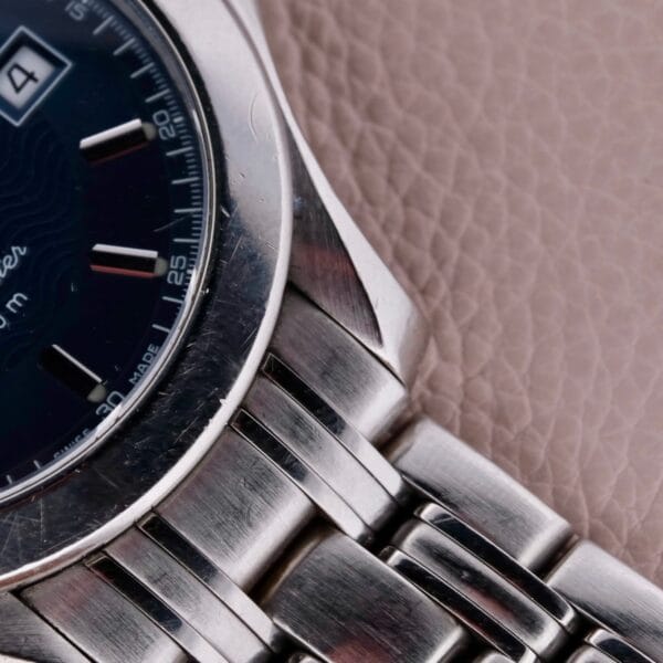 Close-up of a silver wristwatch with a metallic band on a textured surface. The dark watch face displays the brand name Omega Seamaster 120M Blue Wave with the date set to the 4th. Minute markers adorn the dials outer edge, enhancing this sophisticated timeless piece.
