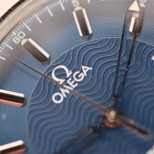 A close-up of the Omega Seamaster 120M Blue Wave watch highlights its blue textured dial, inspired by ocean waves, with silver hour markers and hands, complemented by the brand logo and name.