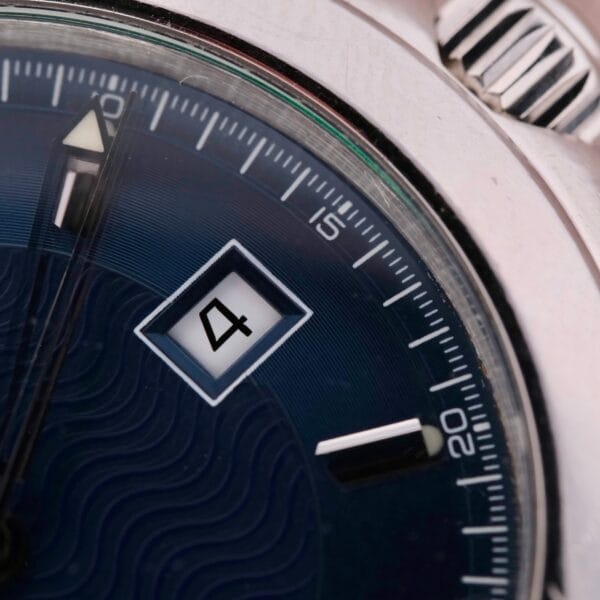 A close-up of the Omega Seamaster 120M Blue Wave wristwatch features a dark blue dial with the number four in a date window. The hands point to 11:07, and the metallic bezel enhances textured dials with a wavy pattern behind the hour markers.