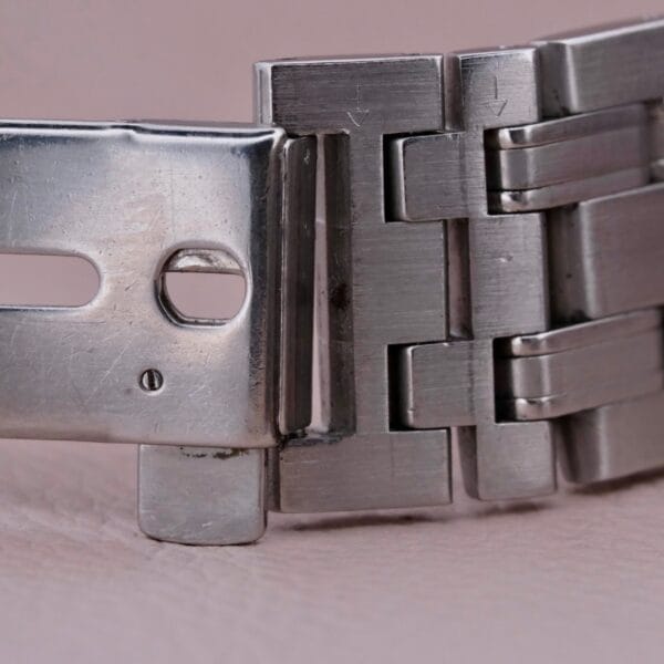Close-up of a metal watch clasp and bracelet with a brushed finish, reminiscent of the Omega Seamaster 120M Blue Wave. The clasp has small holes and a hinge, linking to the bracelet on a beige surface.