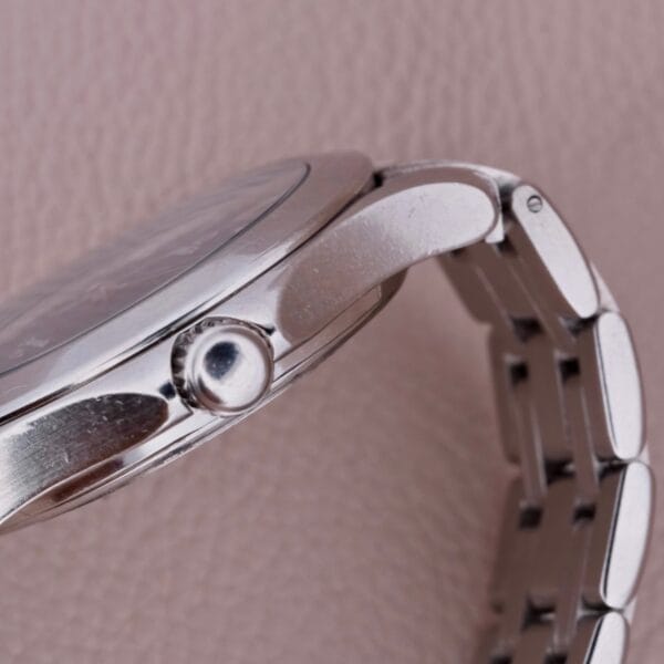 Close-up of an Omega Seamaster 120M Blue Wave wristwatch on a textured beige background, showcasing the side profile with a focus on its round crown for time adjustment. The metal link bracelet elegantly complements this sophisticated timepiece.