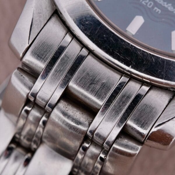 A close-up of a stainless steel band attached to the Omega Seamaster 120M Blue Wave watch reveals brushed metal links with wear marks. The partly visible face adds elegance against a dark background with subtle white highlights.
