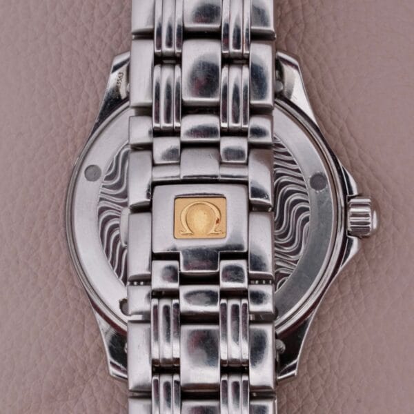 Close-up of a silver Omega Seamaster 120M Blue Wave watch with a detailed metal band and textured backplate featuring a gold square engraved with an omega-like symbol, laid against a taupe leather surface.