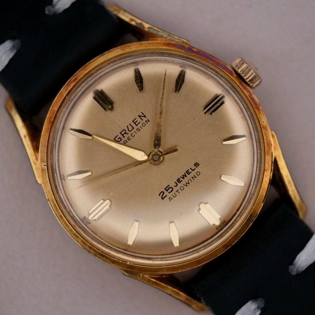 A close-up of the vintage 1960s Gruen Precision 25 Jewels Auto Wind with a gold-toned case features bold black hour markers, two hands, and 25 Jewels Autowind text. It is paired with a classic black strap with white stitching, capturing timeless elegance.