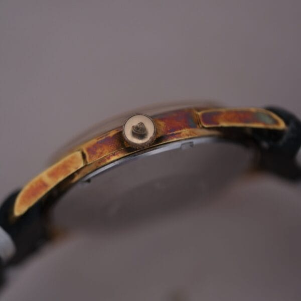 Close-up of a 1960s Gruen Precision 25 Jewels Auto Wind Bombay Lugs with a tarnished metal casing. The timepiece has a small knob on the side and shows wear and aging. The focus is on the side view, highlighting decay on its vintage metal surface.