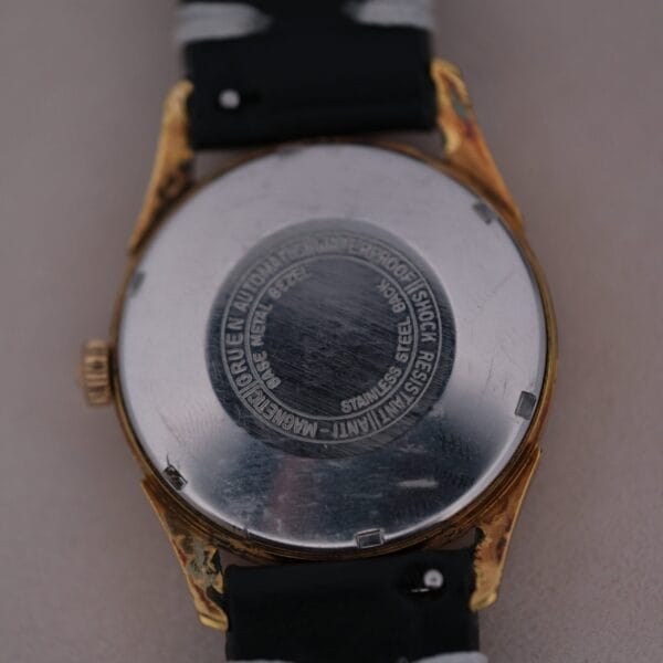 The 1960s Gruen Precision 25 Jewels Auto Wind Bombay Lugs watch showcases its waterproof, shock resist, stainless steel, and antimagnetic features, complemented by a black band with gold-toned edges, embodying the elegance and durability of the era.