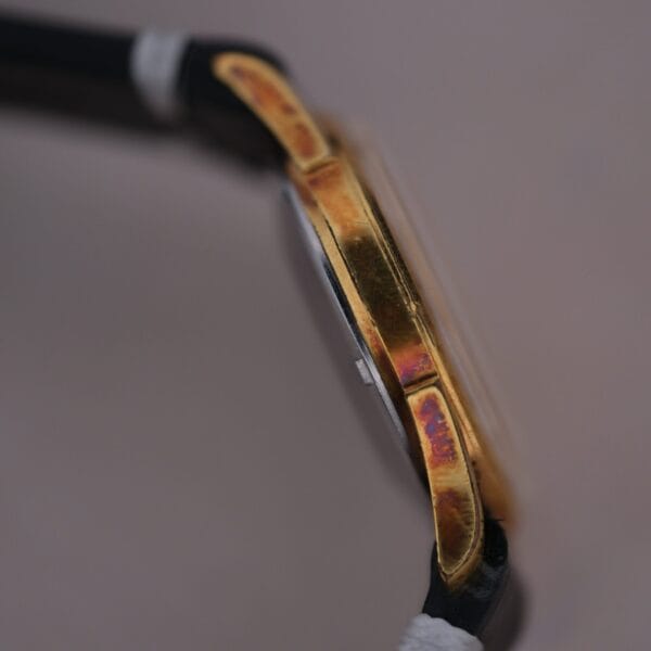 Close-up of a 1960s Gruen Precision 25 Jewels Auto Wind Bombay Lugs watch with a gold-toned rectangular case. The black strap has white ends. The case shows wear and subtle purple-blue discoloration. The background is softly blurred.