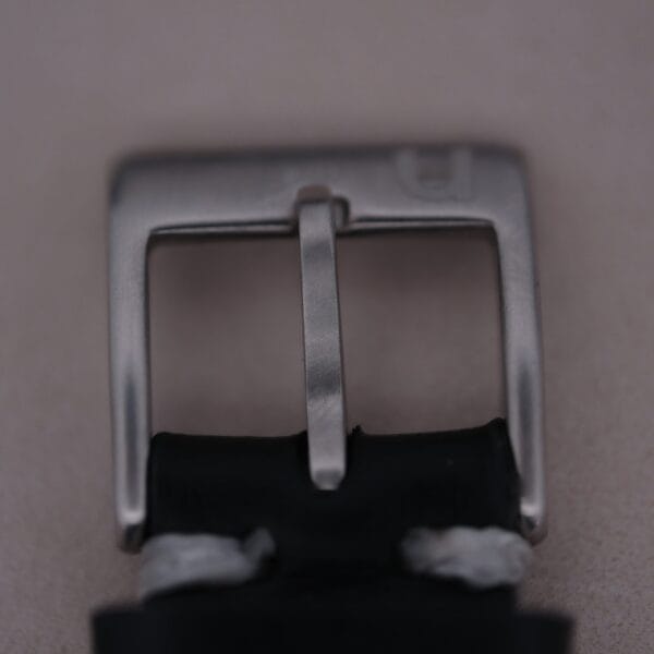 A close-up of a metal belt buckle on a black strap, featuring white edge stitching. Its simple square design is reminiscent of the 1960s Gruen Precision style, akin to the Gruen Precision 25 Jewels Auto Wind Bombay Lugs.