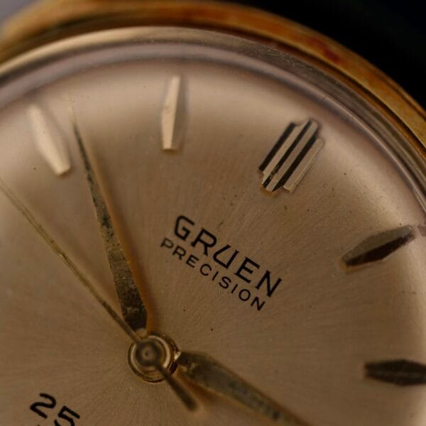 A close-up of the 1960s Gruen Precision 25 Jewels Auto Wind watch reveals gold hour markers and hands. This vintage piece exudes a warm metallic sheen with its minimalist design, small central hand pinion, textured dial, and distinctive Bombay lugs.