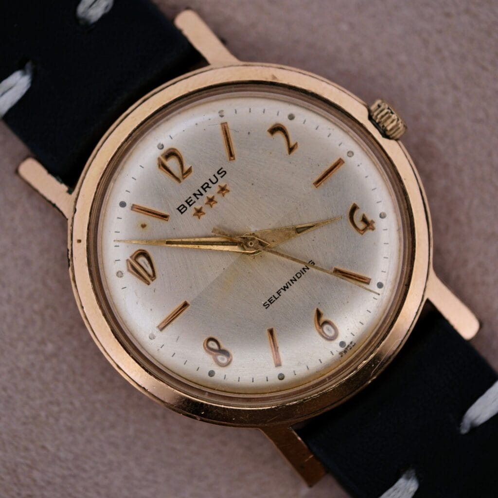 The Benrus 3 Star Automatic Sunburst Dial wristwatch features a gold-tone case marked Selfwinding, a cream dial with gold Arabic numerals and hands, and a black leather strap with white stitching.