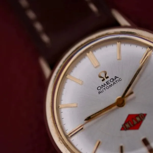 A vintage Omega Automatic Cal. 551 wristwatch for Inland Company showcases a silver dial with gold hour markers, a brown leather strap, and an INLAND logo in red on the face. It rests on a textured maroon background, enhancing its classic elegance.