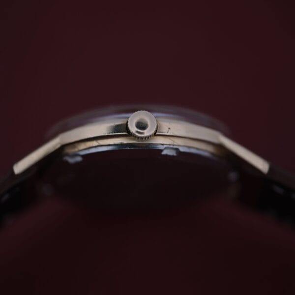 Close-up of an Omega Automatic Cal. 551 for Inland Company, highlighting its thin profile and crown with a metallic case and dark strap, elegantly set against a rich dark brown background.