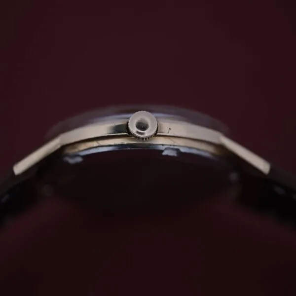 Close-up of an Omega Automatic Cal. 551 for Inland Company, highlighting its thin profile and crown with a metallic case and dark strap, elegantly set against a rich dark brown background.