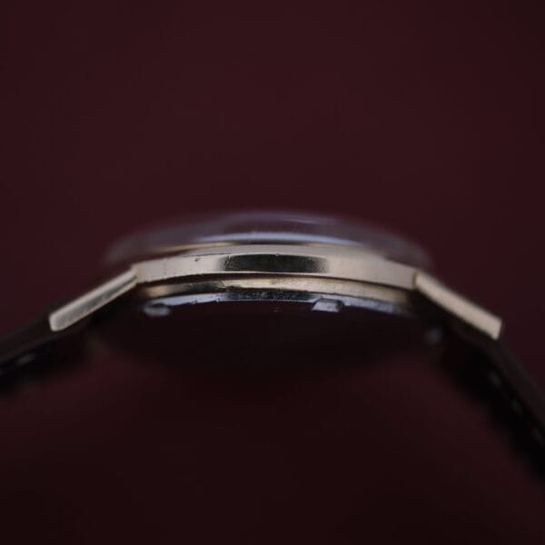 A close-up side view of the Omega Automatic Cal. 551 for Inland Company wristwatch, featuring a thin gold bezel against a dark brown background. The focus is on the edge and thickness, highlighting its sleek design and subtle reflection.