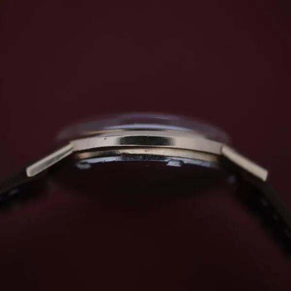 A close-up side view of the Omega Automatic Cal. 551 for Inland Company wristwatch, featuring a thin gold bezel against a dark brown background. The focus is on the edge and thickness, highlighting its sleek design and subtle reflection.