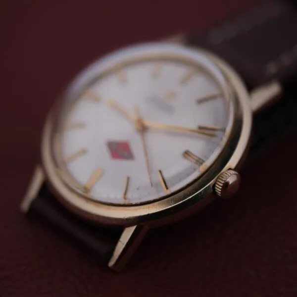 A close-up of the Omega Automatic Cal. 551 for Inland Company wristwatch, featuring a white dial with gold-tone hands and markers, a brown leather strap, and a red rectangular logo near the center. It sits elegantly against a burgundy surface background.