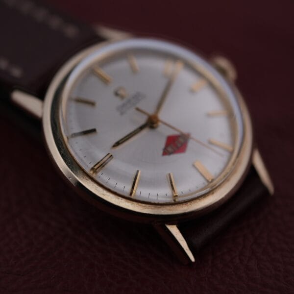A close-up of the Omega Automatic Cal. 551 for Inland Company showcases a white dial, gold markings, and a red emblem. Its dark leather strap elegantly rests on a brown leather surface, exuding timeless sophistication.
