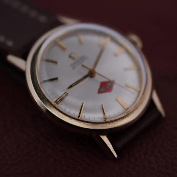 A close-up of the Omega Automatic Cal. 551 for Inland Company showcases a white dial, gold markings, and a red emblem. Its dark leather strap elegantly rests on a brown leather surface, exuding timeless sophistication.