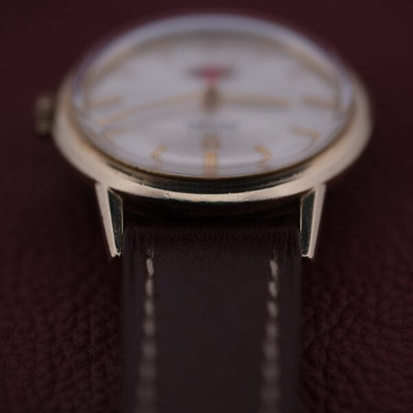 A close-up of an Omega Automatic Cal. 551 for Inland Company wristwatch showcases a golden frame and dark brown leather strap, with a slightly blurred watch face displaying gold hour markers on a rich, textured background.