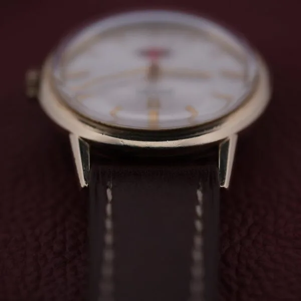 A close-up of an Omega Automatic Cal. 551 for Inland Company wristwatch showcases a golden frame and dark brown leather strap, with a slightly blurred watch face displaying gold hour markers on a rich, textured background.
