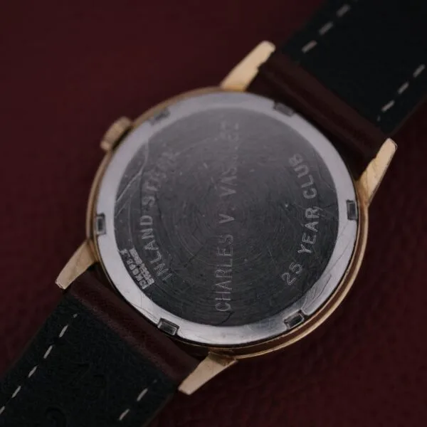 The image shows the back of a watch with INLAND STEEL at the top, CHARLES V. VARRELMAN centered, and 25 YEAR CLUB below. It resembles an Omega Automatic Cal. 551 for Inland Company and features a dark leather strap on a textured surface.