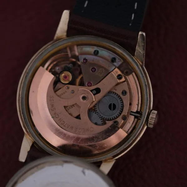 A close-up of the Omega Automatic Cal. 551 for Inland Company displays its intricate gears through an open back. With a metal casing and leather strap, it elegantly rests on a textured dark red surface, highlighting timeless craftsmanship.