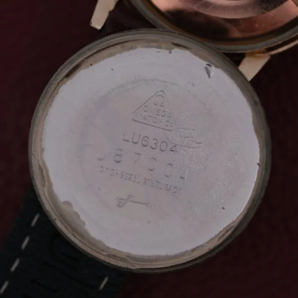 A close-up of a watch case back reveals engravings: Omega Watch Co and model number LU6304, hinting at the precision of the Omega Automatic Cal. 551 for Inland Company. The strap partially shows against a dark red background, evoking timeless elegance on the metallic surface.