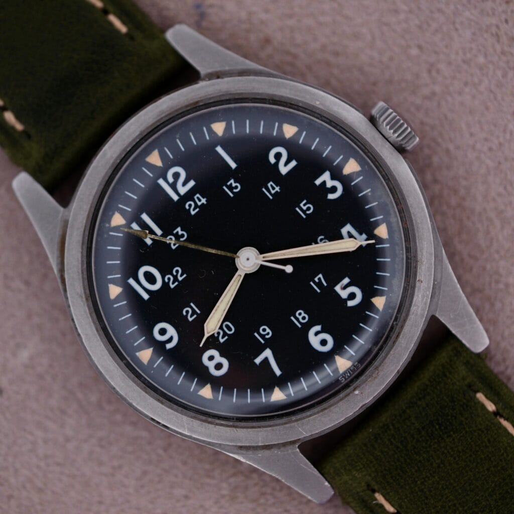 A 1964 Benrus U.S. Military Issued DTU-2A/P Hack Second wristwatch with a black dial, 24-hour markers, and a green fabric strap rests on a beige surface.