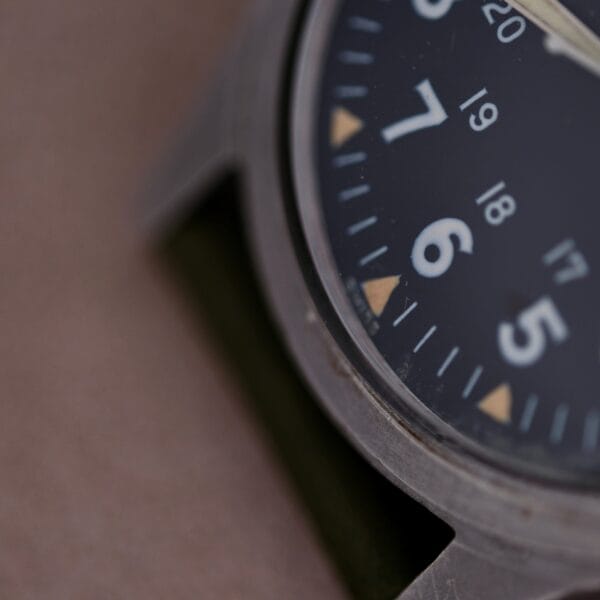 Close-up of a 1964 Benrus DTU-2A/P watch face with large white numerals for 5, 6, and 7 on a dark dial. Smaller numbers 17 to 20 are visible. The Hack Second feature ensures precision, while its metallic casing and dark strap complete the military-issued watch design.