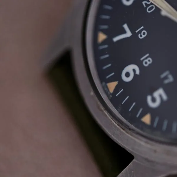 Close-up of a 1964 Benrus DTU-2A/P watch face with large white numerals for 5, 6, and 7 on a dark dial. Smaller numbers 17 to 20 are visible. The Hack Second feature ensures precision, while its metallic casing and dark strap complete the military-issued watch design.