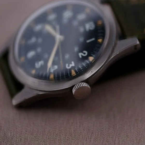 Close-up of a 1964 Benrus U.S. Military Issued DTU-2A/P Hack Second wristwatch with a black dial, white numerals, and stainless steel case. It features a green strap, silver crown, and the time is set against fabric.