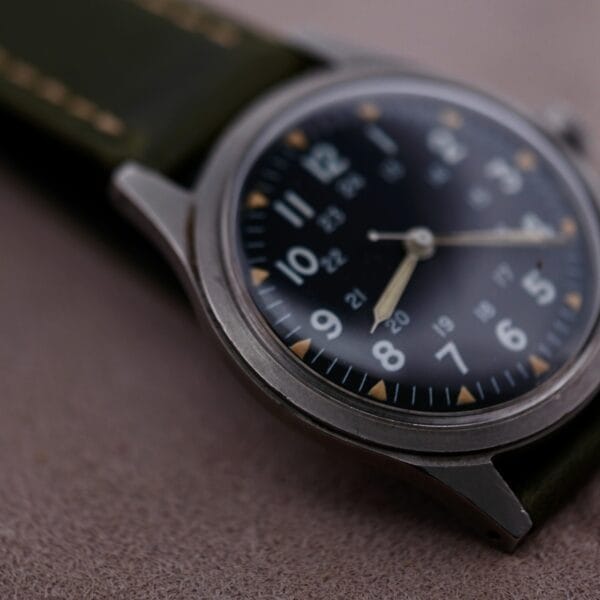 Close-up of the 1964 Benrus U.S. Military Issued DTU-2A/P Hack Second wristwatch, featuring a dark face with white hour numbers, small yellow markers, metallic hands, and a dark green strap. The watch rests on a soft-textured surface.