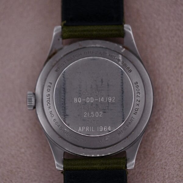 The image shows the back of a 1964 Benrus U.S. Military Issued DTU-2A/P Hack Second wristwatch with engraved numbers, April 1964, and SERIAL NO. Its black strap lies on a soft, textured surface, exuding timeless elegance.