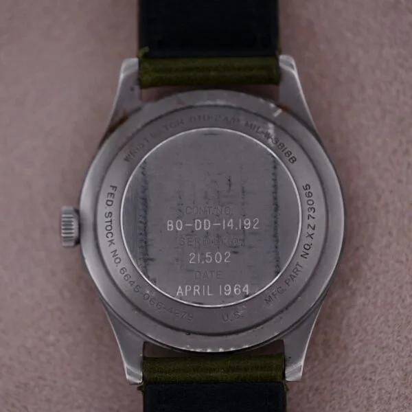 The image shows the back of a 1964 Benrus U.S. Military Issued DTU-2A/P Hack Second wristwatch with engraved numbers, April 1964, and SERIAL NO. Its black strap lies on a soft, textured surface, exuding timeless elegance.