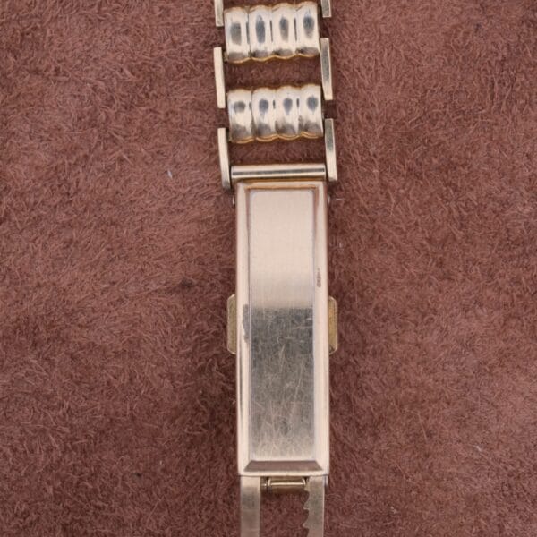 A close-up of an 18k gold Cartier Officer Watch clasp with a textured rectangular surface and link design, evoking exquisite craftsmanship, rests elegantly on a brown fabric background.