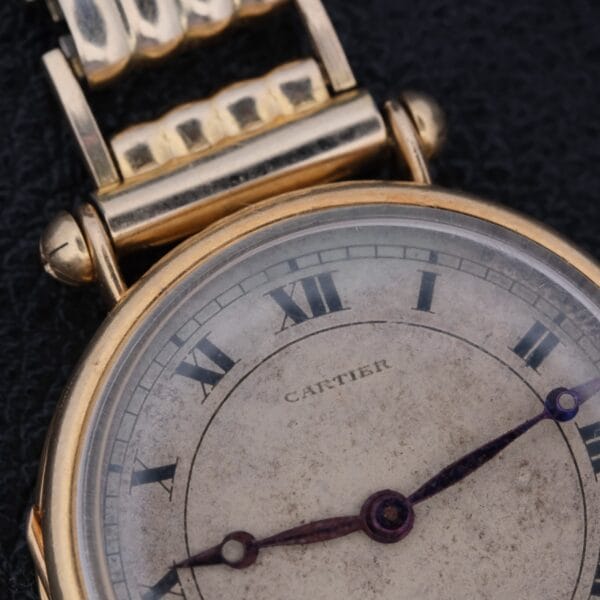 Close-up of the Cartier Officer Watch Edmond Jager, showcasing its Roman numerals and worn 18k gold casing. The delicate hands stand out against a textured dark background.