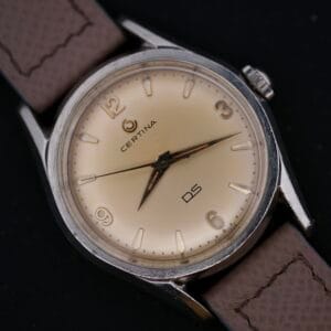 Close-up of a Certina DS 1st Gen Cal. 25-36 in stainless steel with a beige face, brown leather strap, and Arabic numerals at 12, 3, 6, and 9 oclock. Silver hour and minute hands enhance the design against a dark backdrop.