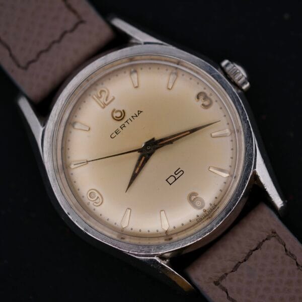 Close-up of a Certina DS 1st Gen Cal. 25-36 in stainless steel with a beige face, brown leather strap, and Arabic numerals at 12, 3, 6, and 9 oclock. Silver hour and minute hands enhance the design against a dark backdrop.