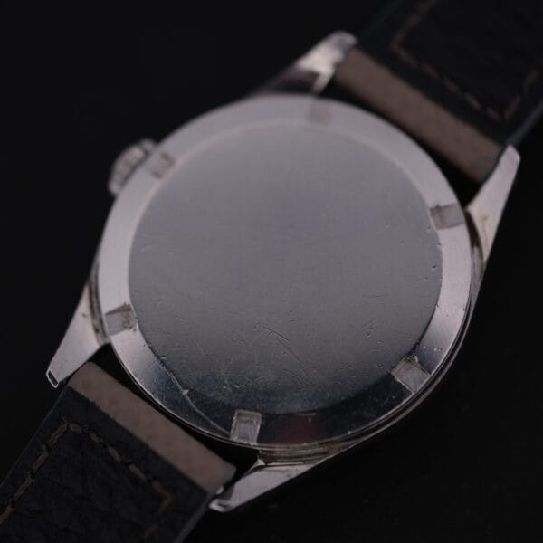 The Certina DS 1st Gen Cal. 25-36 Stainless Steel showcases its character with a brushed metal case back, revealing scratches and marks. A dark leather strap is attached on both sides, while the black background enhances its timeless details.