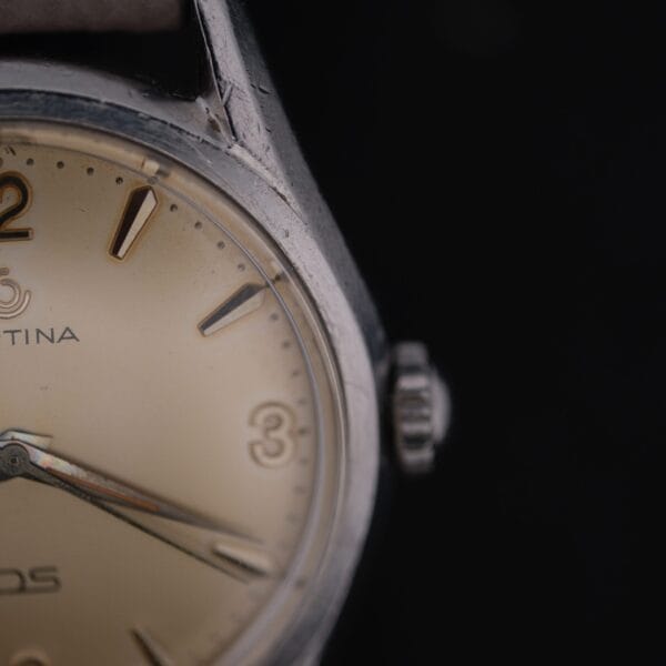 Close-up of a vintage Certina DS 1st Gen Cal. 25-36 wristwatch featuring a beige dial, partial CERTINA name, and silver hour markers. The hands elegantly point to 10:10, with the worn stainless steel case standing out against the black background, emphasizing its classic design.