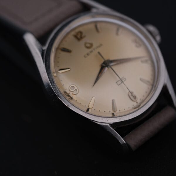 Close-up of a vintage Certina DS 1st Gen Cal. 25-36 wristwatch featuring a beige dial with silver hour and minute hands, dark Arabic numerals, and partially visible branding. The watch includes a brown leather strap and stainless steel case.