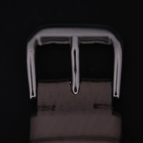 A close-up of a metallic buckle on a dark surface shows a rectangular frame with a single prong, echoing the classic design of the Certina DS 1st Gen Cal. 25-36 Stainless Steel. The partially visible strap below enhances its vintage appeal.
