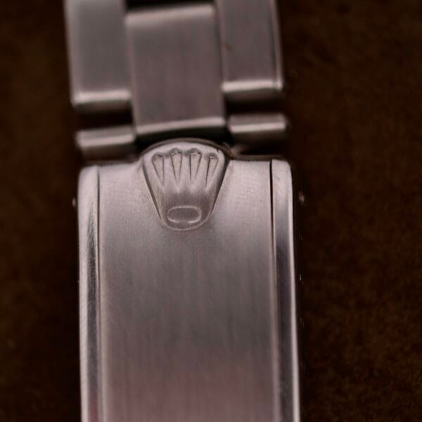 Close-up of a metallic clasp with a raised crown emblem, reminiscent of the Rolex Oyster Perpetual Datejust 6605 Cal. 1066, featuring a brushed texture and extending links, set against a dark background.